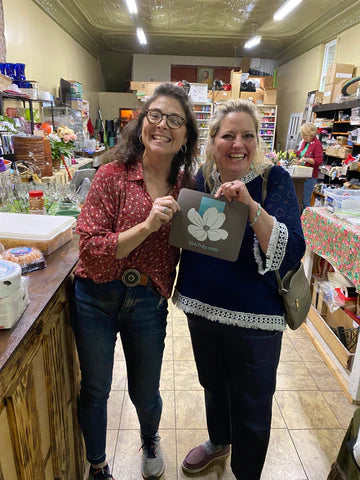 Two women are smiling while holding a card with UrnTray's logo on it