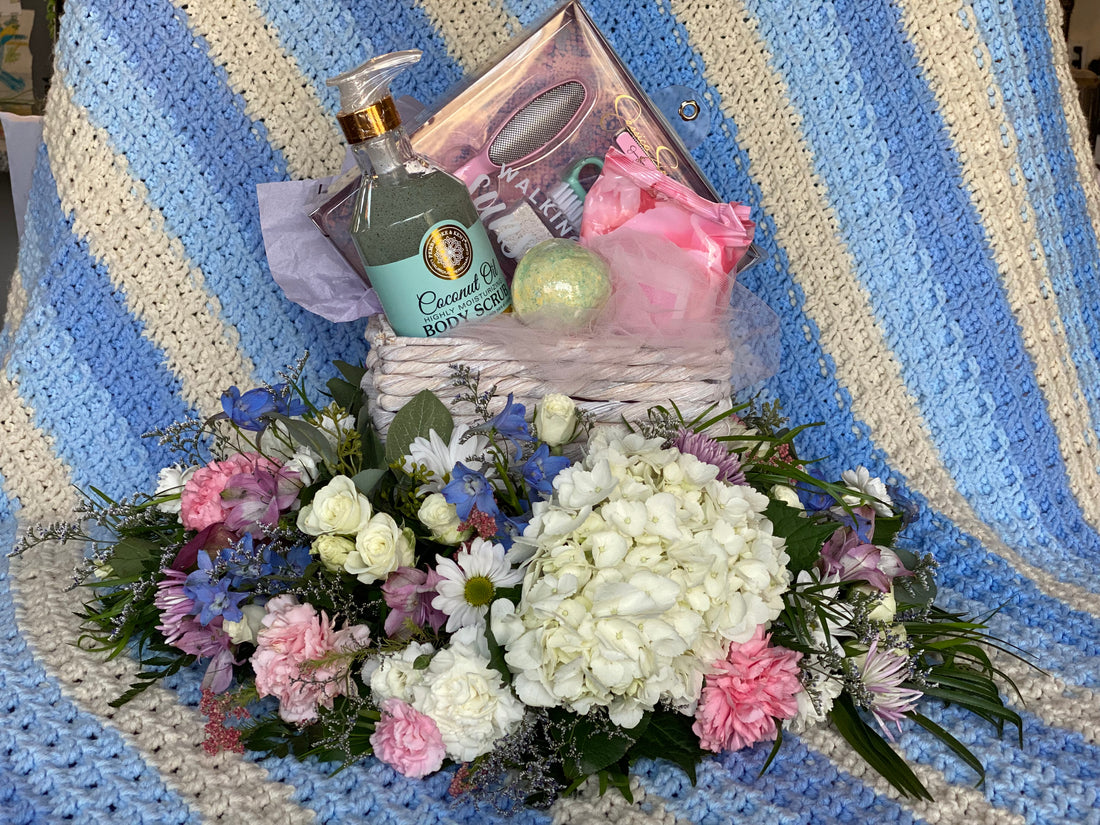 A gift basket filled with pampering items sets on the platform of an UrnTray designed with pink, white, and blue flowers.