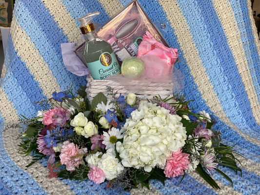 A gift basket filled with pampering items sets on the platform of an UrnTray designed with pink, white, and blue flowers.