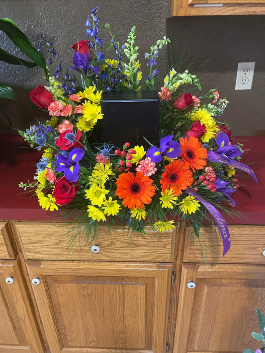 A black urn sits on a floral wreath platform tray, with happy flowers surrounding.