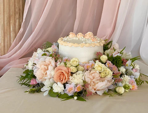 A white and peach wedding cake sits on the pedestal of an UrnTray with peach and white flowers surrounding the base.