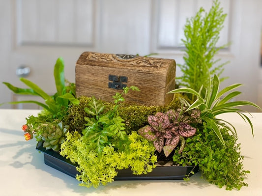 A small wooden treasure chest sets on the pedestal of an UrnTray with a variety of live plants surrounding. 
