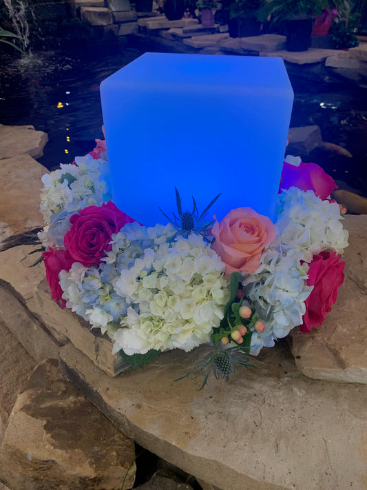 A large glass cube sits on a floral wreath tray by UrnTray. The wreath has white and pink flowers, the cube is illuminated with blue light.