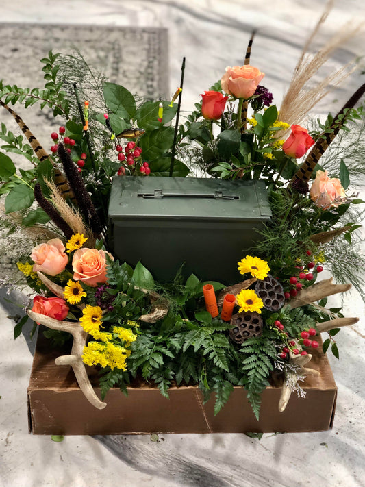 An urn wreath with peach roses, red hypericum berries, yellow daisies, lotus pods, antlers, pampas grass, shot gun shells, and pheasant feathers surrounds an urn that looks like an old ammo case.